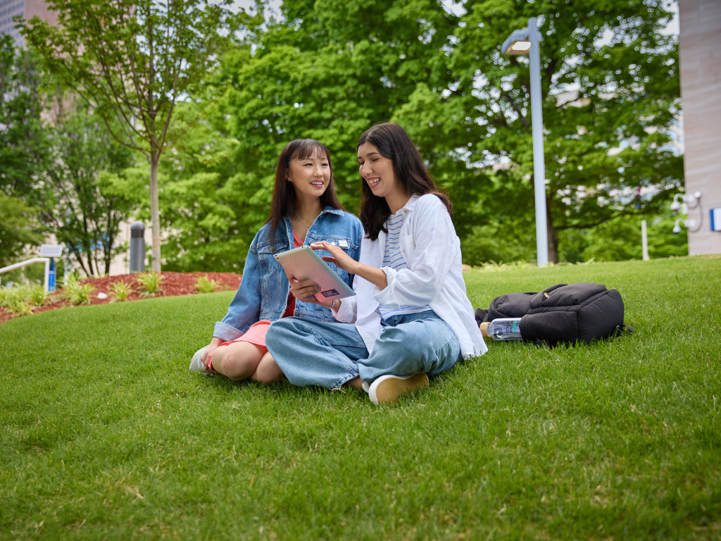 Software engineering students sit in a park on the grass. They are smiling and reviewing content on a tablet. 