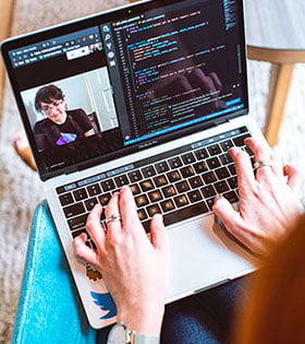 Person's hands typing on a laptop with a video meeting on one side of the screen and code on the other