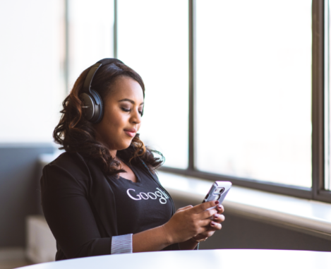 Woman of color wearing google shift and blazer, wearing headphones and looking at cell phone