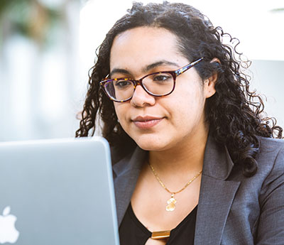 Woman of color wearing glasses and a blazer working in front of an Apple laptop